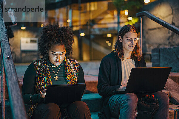 Junge männliche und weibliche Studenten  die einen Laptop benutzen  während sie nachts auf Stufen auf dem Campus sitzen