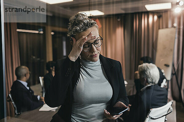 Besorgte reife Geschäftsfrau  die ihre Stirn berührt  während sie auf ihr Smartphone schaut  gegenüber Kollegen im Sitzungssaal
