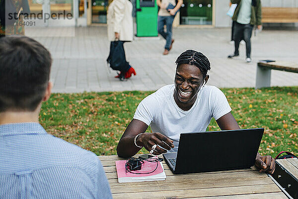 Junger männlicher Student lachend beim Sitzen mit Freund in Campus
