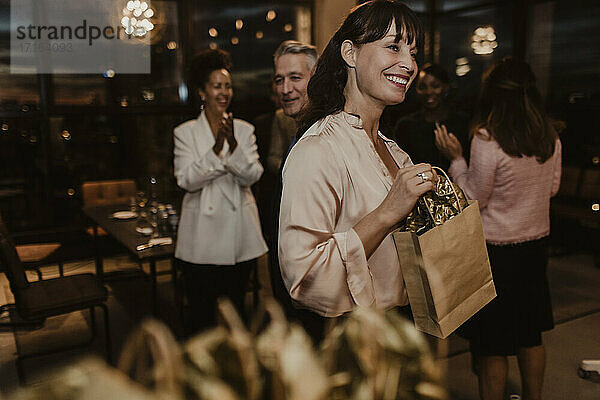 Lächelnde Unternehmerin mit Geschenktüte  die während einer Firmenfeier wegschaut