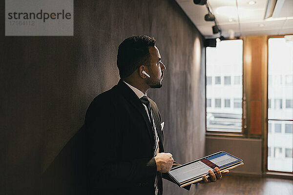 Nachdenklich männlichen Profi hält digitale Tablette Blick weg  während gegen die Wand im Sitzungssaal im Büro lehnt