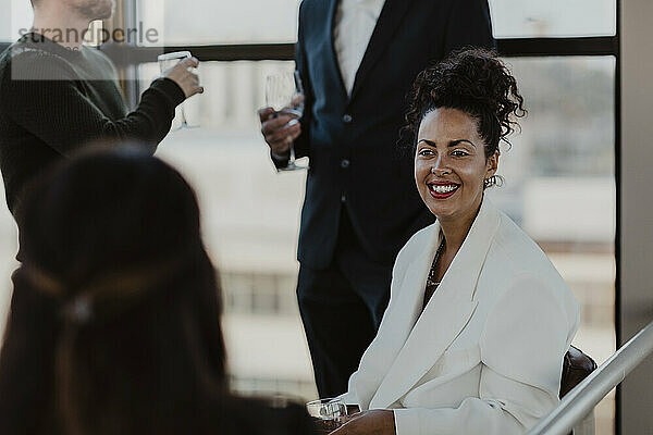 Lächelnde Geschäftsfrau mit Weinglas im Gespräch mit Kollegen während einer Firmenfeier