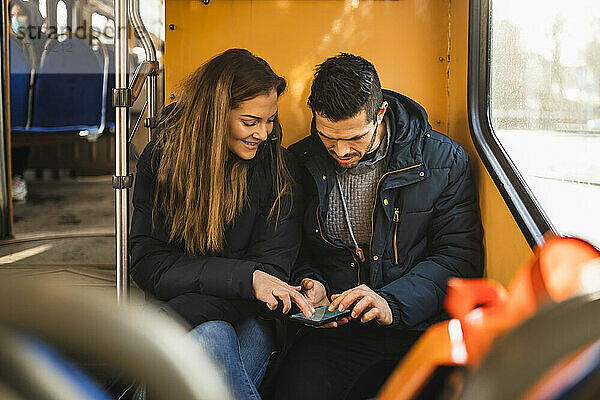 Heterosexuelles Paar  das während der Pandemie in der Straßenbahn sitzt und ein Smartphone benutzt