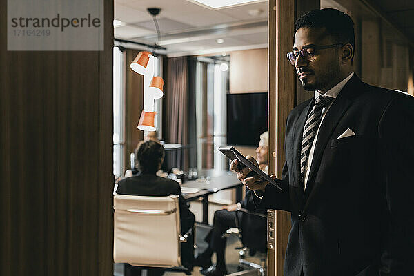 Männlicher Unternehmer  der ein Smartphone benutzt  während er sich im Büro an die Tür zum Vorstandszimmer lehnt