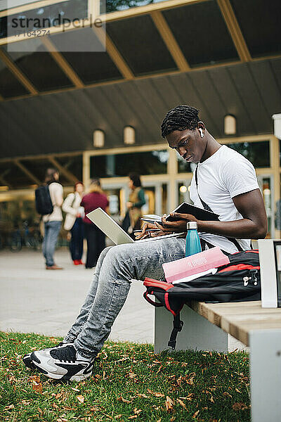 Afrikanischer männlicher Student  der ein Buch liest  während er auf einer Bank auf dem Universitätscampus sitzt