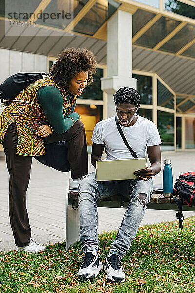 Männlicher Student beim E-Learning am Laptop  während seine Freundin auf dem Campus steht