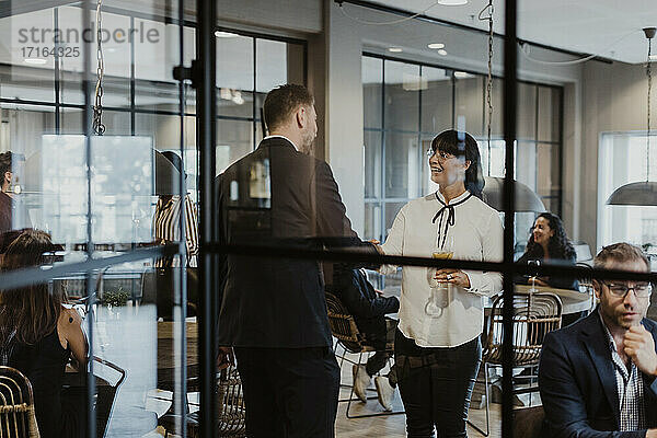 Geschäftsfrau schüttelt Hände mit männlichen Kollegen durch Glas während gesellschaftlicher Veranstaltung im Büro gesehen