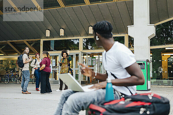 Männlicher Student  der seine Freunde anschaut  während er einen Laptop auf dem Campus benutzt