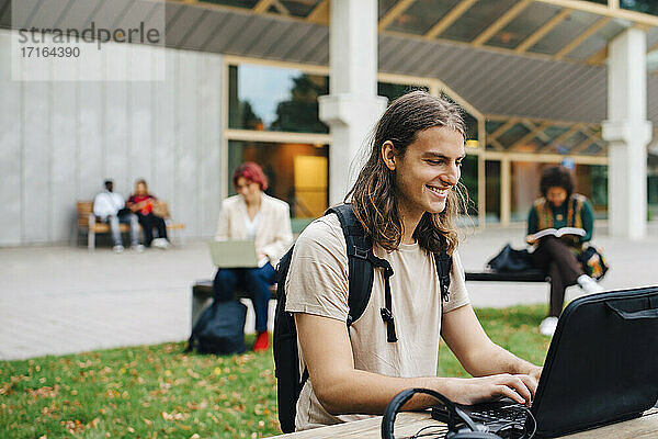 Lächelnder männlicher Student beim E-Learning mit Laptop auf dem Campus