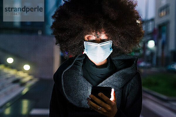 Frau mit Gesichtsmaske  die auf ihr Mobiltelefon schaut  nachts beleuchtet