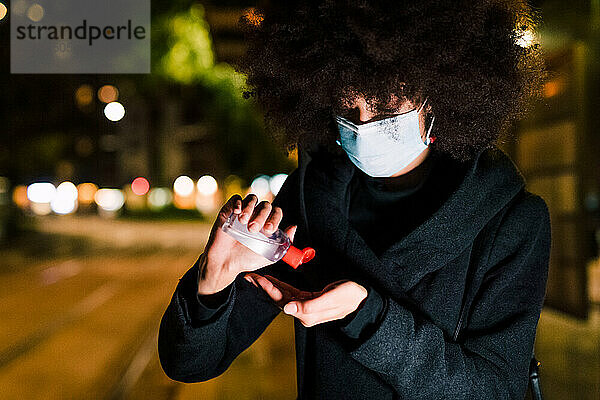 Junge Frau trägt eine Gesichtsmaske und trägt Handdesinfektionsmittel auf  nachts im Freien