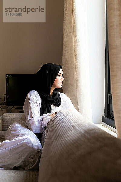 Junge muslimische Frau schaut durch das Fenster zu Hause
