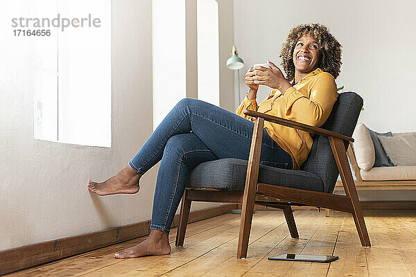 Lächelnde Frau mit Teetasse  die wegschaut  während sie zu Hause auf einem Sessel sitzt