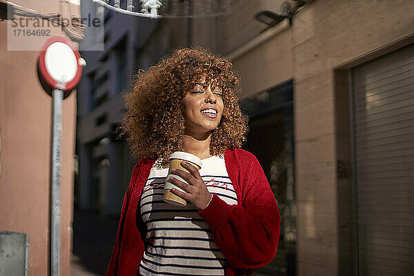 Beautiful woman with disposable coffee cup smiling while standing against street