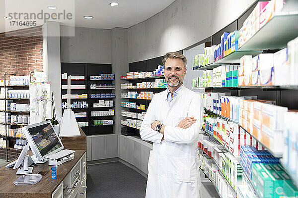 Male pharmacist with arms crossed standing by shelf in store
