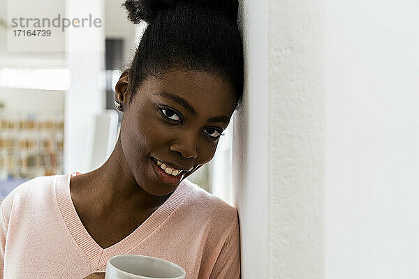 Lächelnde Frau mit Kaffeetasse starrt  während lehnt in der Wand zu Hause