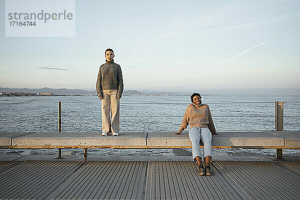Traurige Frau steht in der Ferne mit einem glücklichen Freund  der auf einer Bank am Meer sitzt