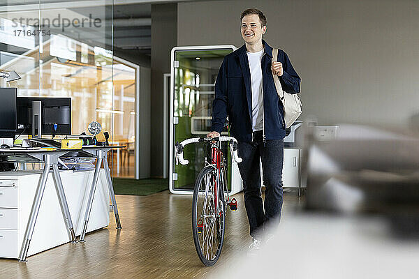 Lächelnder Geschäftsmann mit Tasche und Fahrrad  der nach der Arbeit aus dem Büro geht