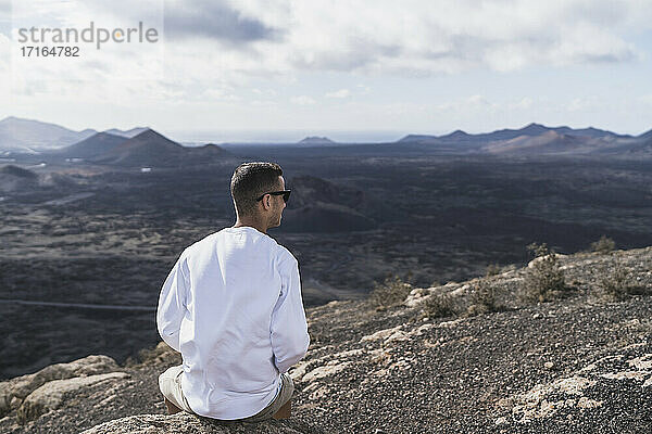 Männlicher Tourist  der auf einem Felsen sitzt und die Aussicht auf den Vulkan El Cuervo an einem sonnigen Tag betrachtet  Lanzarote  Spanien