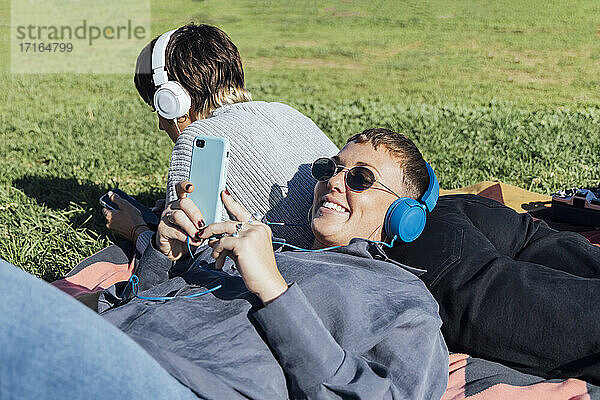 Lächelnde Frau mit Kopfhörern  die ein Mobiltelefon benutzt  während sie sich auf einen im Park liegenden Freund stützt