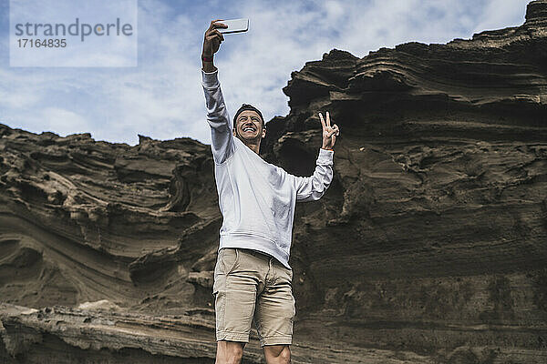Lächelnder männlicher Tourist  der Frieden gestikuliert  während er ein Selfie vor einem Felsen in El golfo  Lanzarote  Spanien macht
