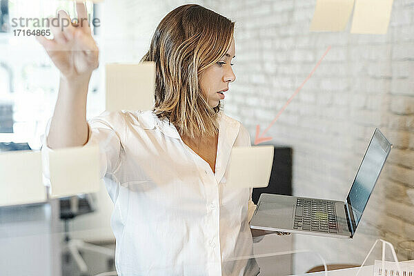 Geschäftsfrau  die einen Laptop hält und eine Geschäftsstrategie im Büro vorbereitet  gesehen durch Glas