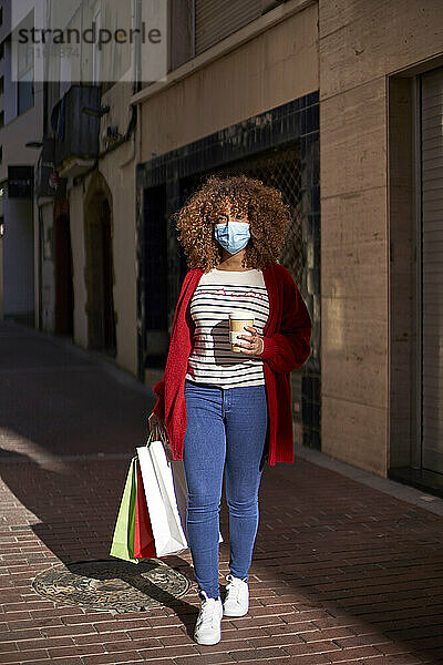 Young woman wearing protective face mask holding coffee cup and shopping bag while walking on footpath