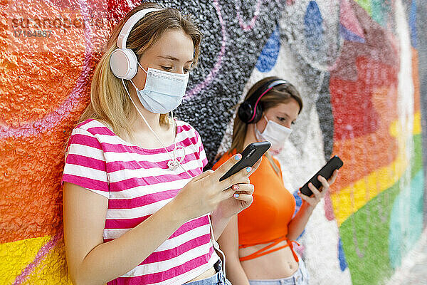 Freundinnen  die ein Mobiltelefon benutzen  während sie an einer bunten Wand Musik hören  während COVID-19