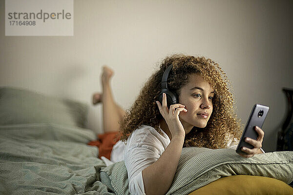 Junge Frau benutzt ihr Smartphone und hört Musik  während sie auf dem Bett liegt