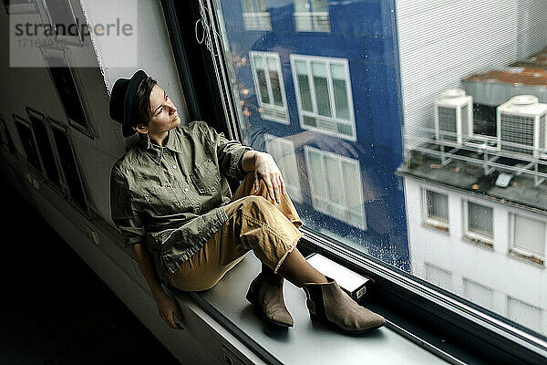Modische Frau  die durch ein Fenster schaut  während sie auf dem Fensterbrett eines Ateliers sitzt