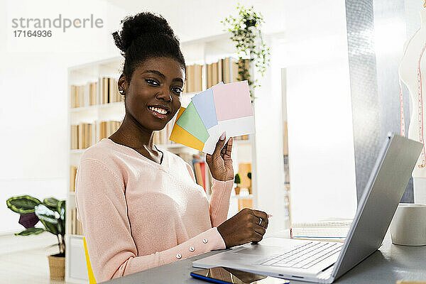 Lächelnde Modestylistin mit Laptop  die bunte Stoffmuster zeigt  während sie zu Hause im Büro sitzt
