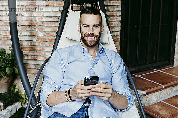 Lächelnder junger Mann  der mit seinem Handy arbeitet  während er in der Hängematte auf der Terrasse sitzt