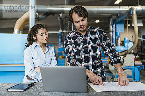Young businessman with document and laptop working while standing by colleague at industry