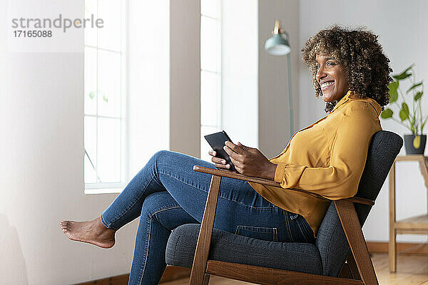Mittlere erwachsene Frau mit digitalem Tablet  die wegschaut  während sie zu Hause auf einem Sessel sitzt