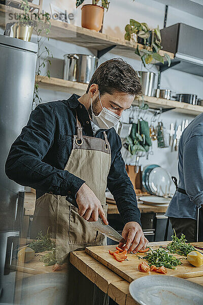 Mid adult chef wearing protective face mask cutting vegetable while standing with colleague in kitchen