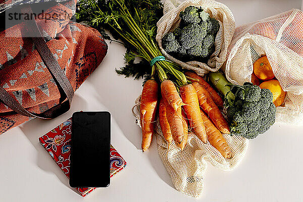 Smartphone und Bio-Gemüse in wiederverwendbaren Netzbeuteln auf dem Küchentisch
