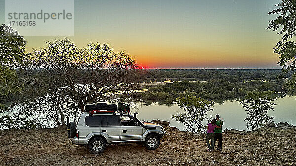 Luftaufnahme eines Paares mit ihrem Jeep  das den Sonnenuntergang vor dem Fluss genießt  Cunene-Flussgebiet  Angola