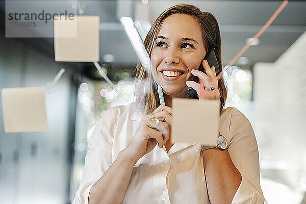 Nahaufnahme einer lächelnden Geschäftsfrau  die über ein Smartphone im Büro spricht  gesehen durch eine Glaswand
