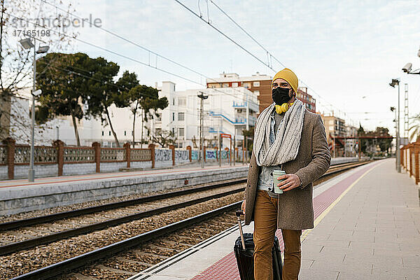 Mann mit Schutzmaske hält Kaffeetasse und Gepäck beim Gehen auf dem Bahnhof