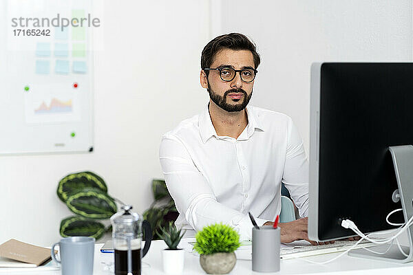 Junger Geschäftsmann mit Brille sitzt vor einem Computer auf einem Schreibtisch am Arbeitsplatz