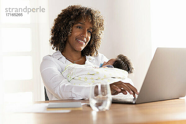 Lächelnde Mutter mit Baby sitzend bei der Arbeit am Laptop zu Hause