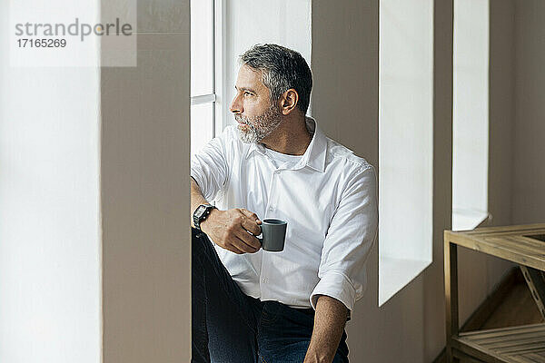 Nachdenklicher Geschäftsmann mit Kaffeetasse  der durch das Fenster schaut  während er zu Hause sitzt