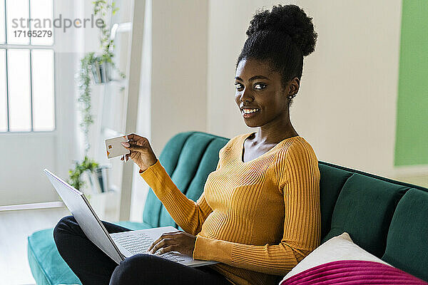 Smiling young Afro woman holding credit card and laptop for online shopping at home
