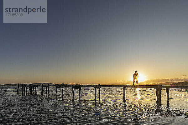 Silhouette einer Frau  die bei Sonnenaufgang allein auf einem Steg an der Küste steht