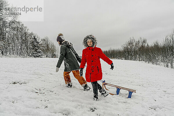 Teenager-Mädchen  das einen Schlitten zieht  während ein kleines Mädchen im Winter im Schnee läuft