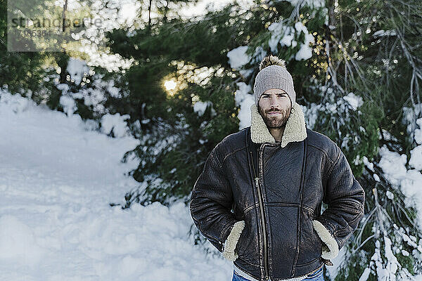 Junger Mann mit Händen in den Hosentaschen  der gegen Bäume im Schnee steht
