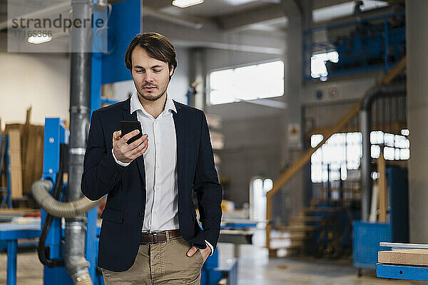 Young businessman using mobile phone while standing with hands in pockets at industry