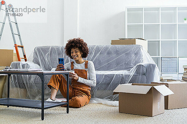 Lächelnde junge Frau  die ein Mobiltelefon benutzt und eine Kaffeetasse in einer neuen Wohnung hält