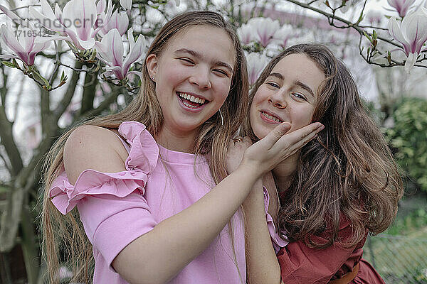 Cheerful female friends embracing against magnolia tree in public park