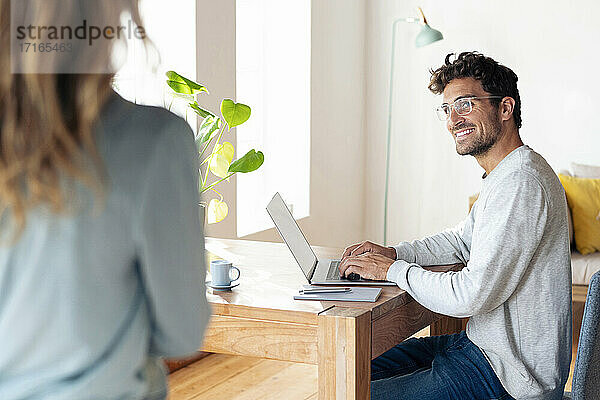 Lächelnder Mann  der eine Frau ansieht  während er im Büro zu Hause am Laptop arbeitet